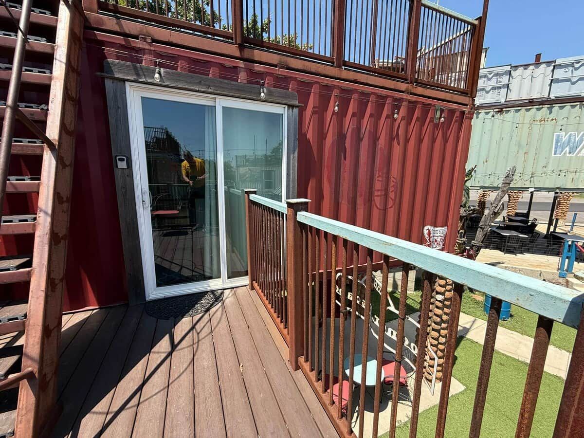 A wooden deck with a glass door leading to a covered area, a metal railing, and a green container in the background.