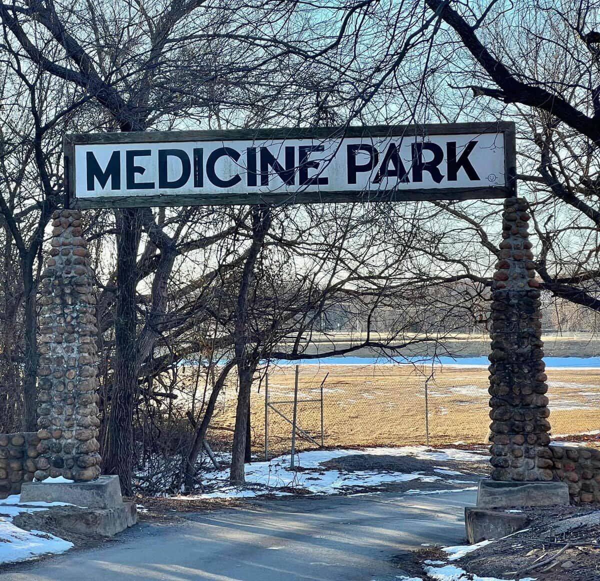 Een bord voor Medicine Park met stenen pilaren en een pad dat door een natuurlandschap loopt.