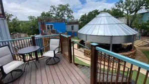 Eine Holzterrasse mit zwei Stühlen und einem Tisch, ein Metallpavillon und ein blaues Gebäude im Hintergrund.
