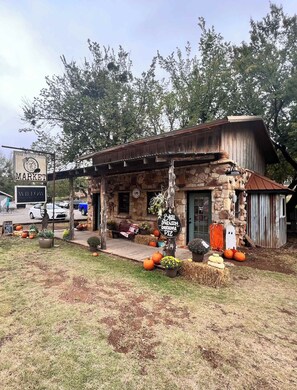 Un edificio rústico de piedra con techo de metal, un cartel de mercado y calabazas y pacas de heno en el césped.