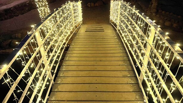 Eine hölzerne Brücke mit Geländern, die mit Lichtern verziert sind, führt zu einem Steinweg.
