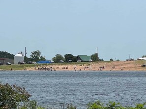 A lakeside area with a sandy beach, a large white tank, and a few buildings in the background. - Sooner Escape: Wichita Mtns Tiny House Stay w/rooftop deck (Medicine Park)