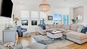 A modern living room with a large sectional sofa, a coffee table, and a chandelier. There are large windows with blinds, a flat-screen TV mounted on the wall, and a small side table with a lamp. - Luxury Lagoon Front Retreat | Private Pool | Pier and Beach Access (Gulf Shores)