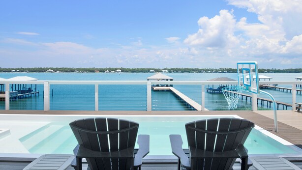 A poolside area with two Adirondack chairs facing a water body, a basketball hoop, and a dock extending into the water. - Luxury Lagoon Front Retreat | Private Pool | Pier and Beach Access (Gulf Shores)