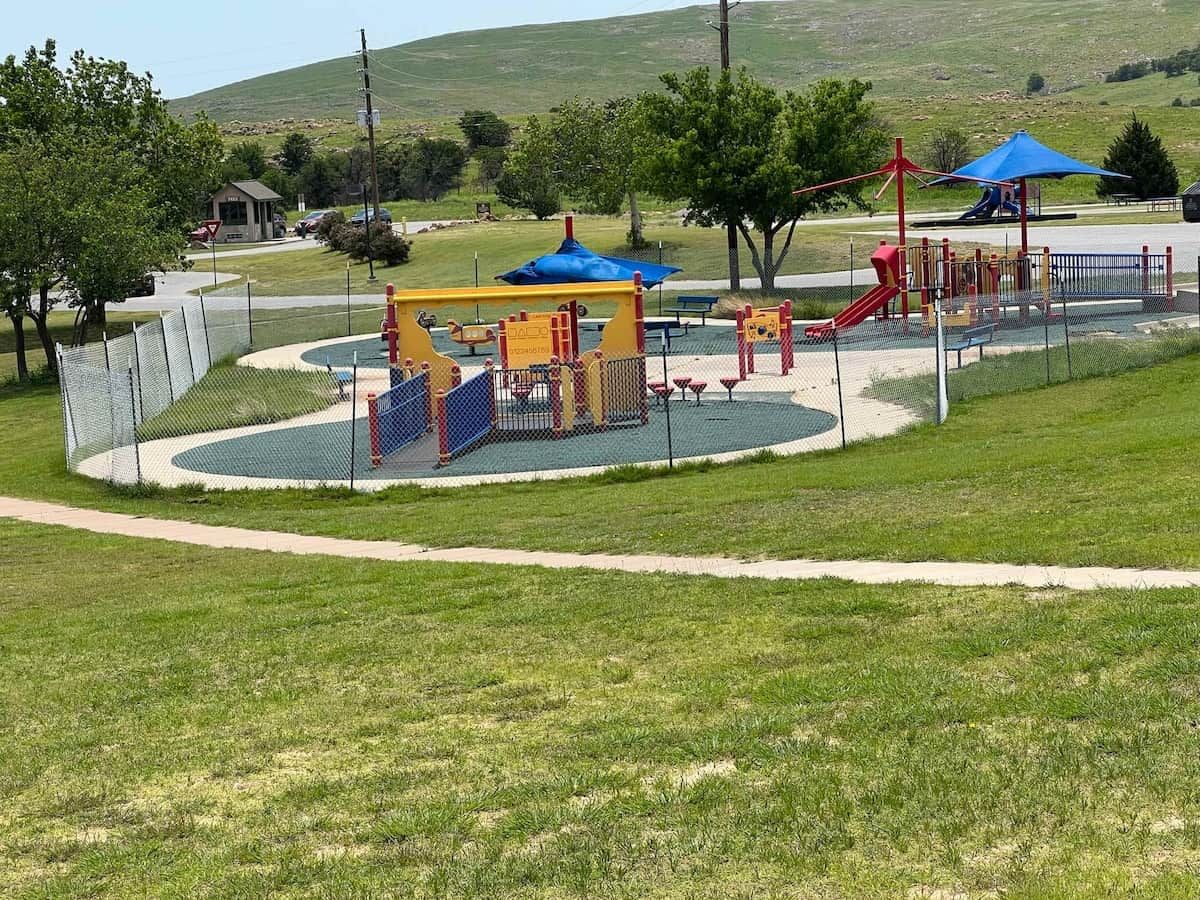 A playground with various play structures, surrounded by a fenced area and grassy fields.