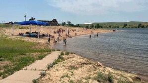 Una playa junto al lago con gente nadando y tomando el sol, un paseo marítimo de concreto y una zona verde con un toldo azul.