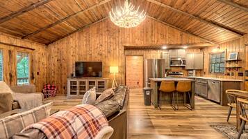 A cozy cabin interior with a wooden ceiling, a television, a sofa, a dining area, and a kitchen.