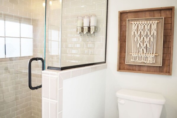 A bathroom with a glass shower enclosure, a white toilet, and a macrame wall hanging. - Historic Midtown Ruidoso Cabin | Walk to Shops & Private Hot Tub (Ruidoso)