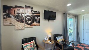 Habitación de hotel con piso a cuadros, dos sillones de cuero negro, una pequeña mesa redonda con lámpara, un televisor montado en la pared y una gran obra de arte con temática de motocicletas sobre los sillones.