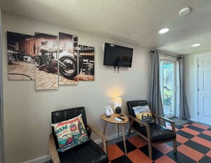 Hotelkamer met een zwart-wit geruite vloer, twee zwarte leren fauteuils, een kleine ronde tafel met lamp, een televisie aan de muur en een groot kunstwerk met motorfietsmotieven erboven.