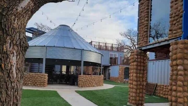 Un espacio al aire libre techado con techo metálico, rodeado de pilares de piedra y un césped con un sendero que lleva a un edificio.