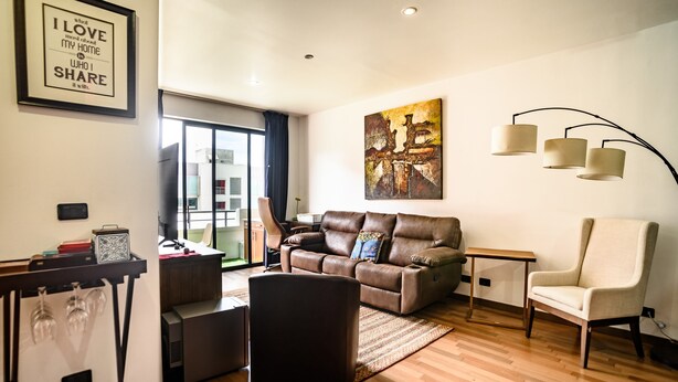 Ein modernes Wohnzimmer mit einem Ledersofa, einem weißen Sessel, einem Couchtisch und einem Beistelltisch. An der Wand hängt ein gerahmter Spruch und ein Gemälde über dem Sofa.