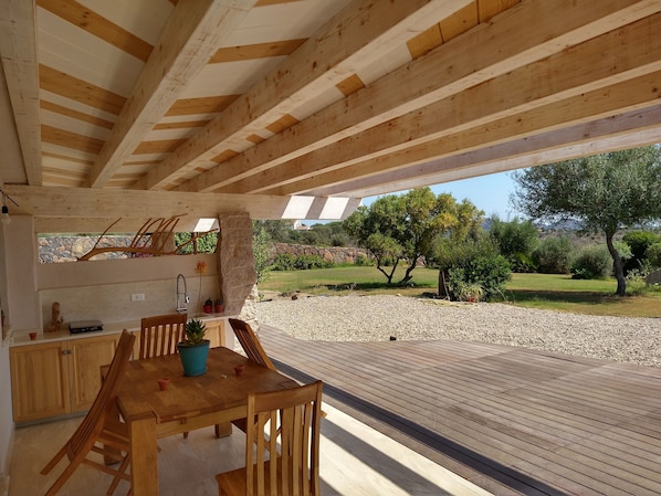 A wooden deck with a table and chairs, a stone pillar, and a view of a landscaped garden with trees and a stone wall. - Villa Giorgia – Sardinia, Orosei: An Entire House with Views of the Valley and the Sea (Orosei)