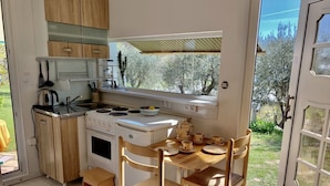 Een kleine keuken met een fornuis, gootsteen en houten kastjes. Er staat een eettafel met stoelen en een raam met uitzicht op een tuin.