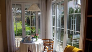Ein Essbereich mit Tisch für zwei Personen, einem Stuhl, einer gelben Decke und Blick auf einen Garten durch große Glasüren.