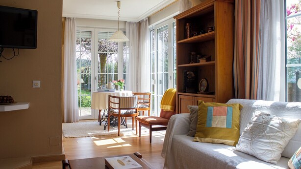 Ein gemütliches Wohnzimmer mit einer Couch, einem Esstisch und Stühlen. Ein Bücherregal und ein an der Wand befestigter Fernseher sind ebenfalls vorhanden.