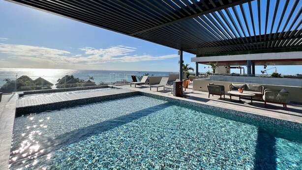 A modern outdoor swimming pool with a view of the ocean, lounge chairs, and a covered seating area. - 1BR Condo Best Location (Playa del Carmen)