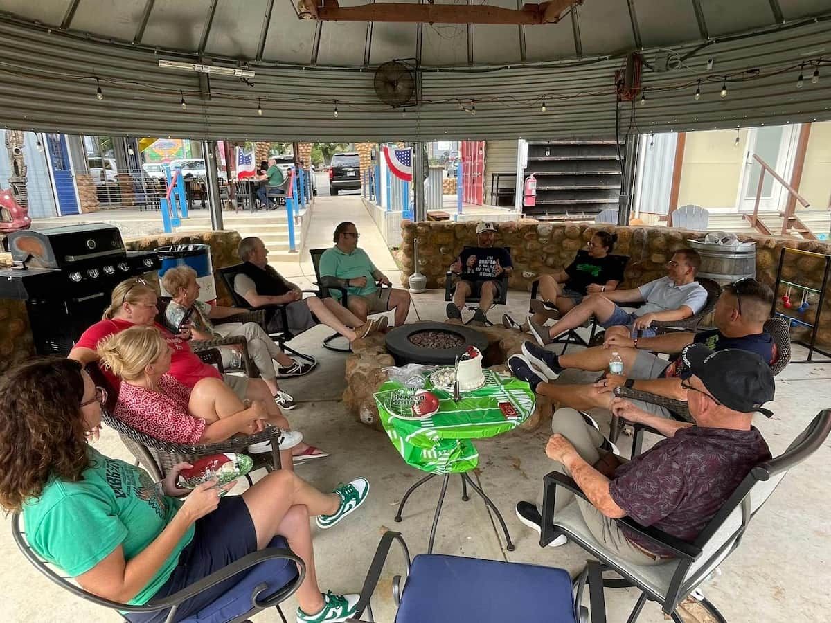 Um grupo de pessoas sentado à mesa, com bolo e bebidas, sob uma área externa coberta e vista para a rua.
