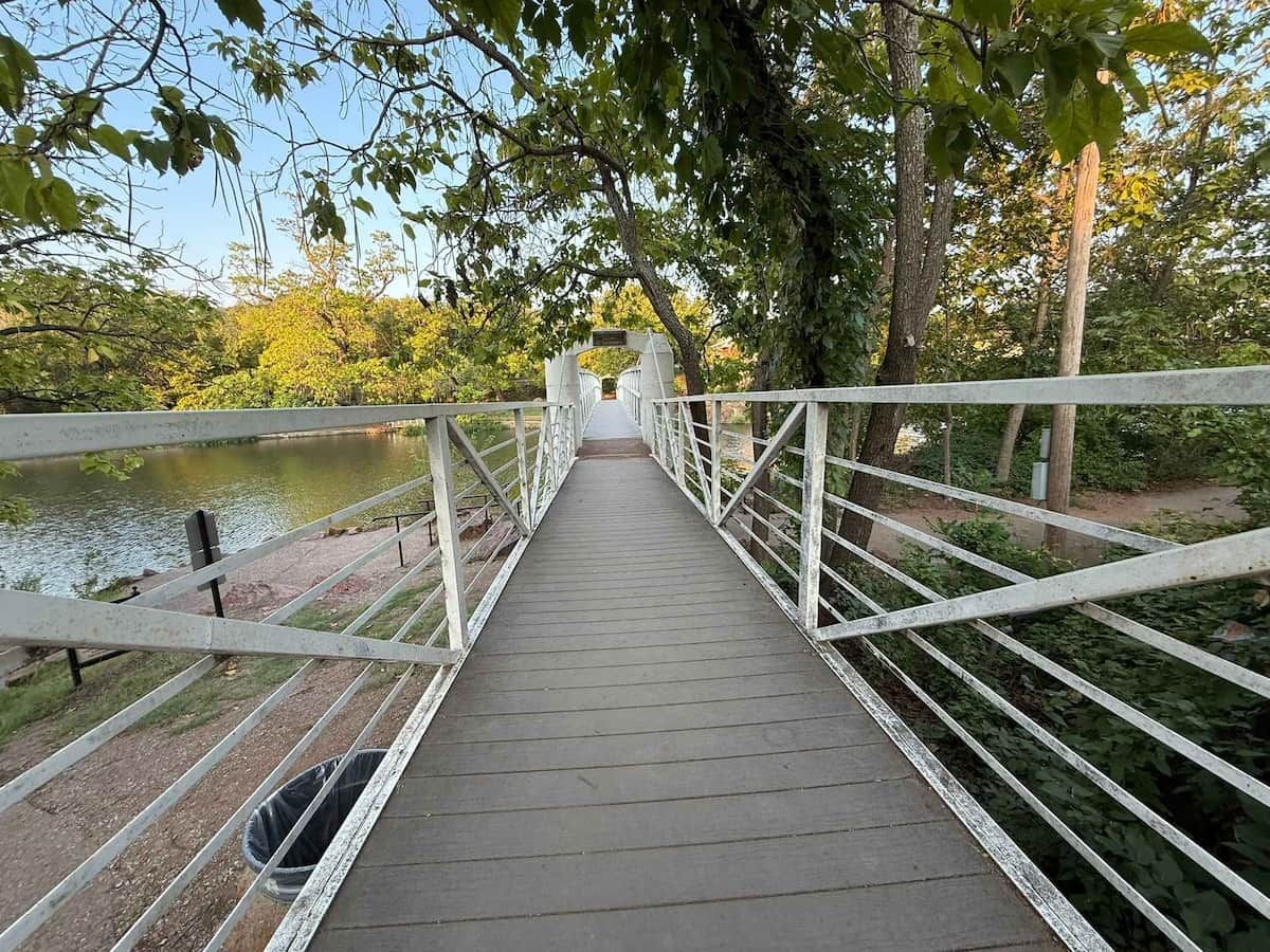 A wooden pedestrian bridge with white railings, leading over a body of water, surrounded by trees.