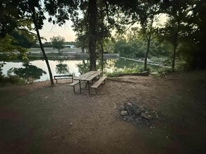 Un espace de pique-nique équipé d’un banc et d’une table à pique-nique, entouré d’arbres et bordé par un plan d’eau.