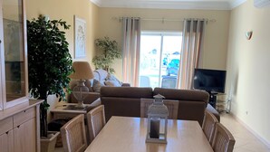 A dining area with a wooden table and wicker chairs, a sofa, a TV, and a large potted plant.