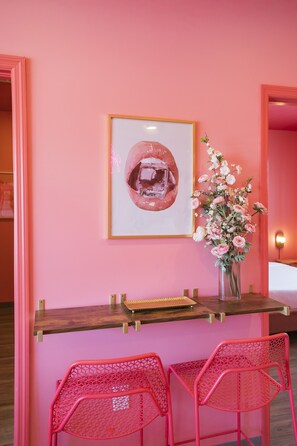 Quarto em tons de rosa, com escrivaninha de madeira, duas cadeiras de rede vermelhas, uma obra de arte emoldurada e um vaso com flores.