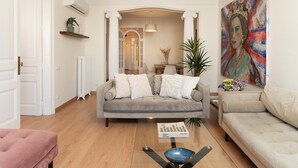 A living room with a grey sofa, a pink ottoman, and a coffee table with a bowl. There is a painting on the wall and a plant on the side table.