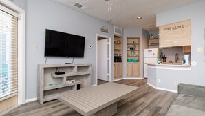 A modern living room with a flat-screen TV, wooden shelves, a bench, and a kitchen area with wooden cabinets and a countertop. - Inviting Coastal Stay Near Beach With Pool, Hot Tub, and Private Balcony (Corpus Christi)