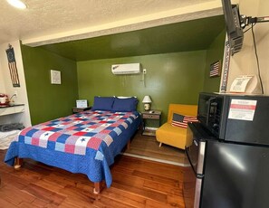 A room with a bed, a nightstand, a lamp, a refrigerator, and a small table with an American flag.