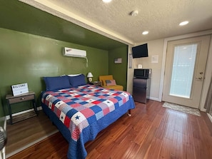 Un dormitorio con cama, mesita de noche, televisor, refrigerador y puerta con ventanal.