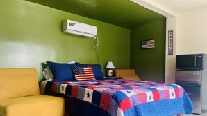 Una habitación con una cama, un sillón amarillo, un kitchenette y una pared verde.