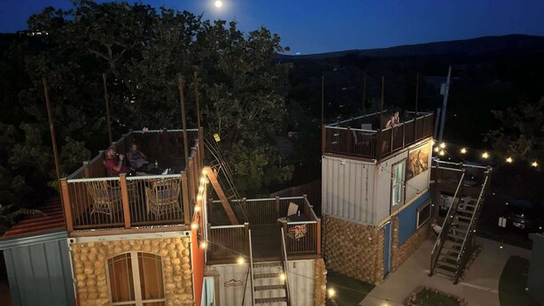Estructura de cabaña única, de varios niveles, con fachada de piedra, terraza de madera y zona de estar al aire libre.