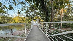 Un puente peatonal de madera con barandillas blancas que conduce a un sendero rodeado de árboles y una masa de agua.
