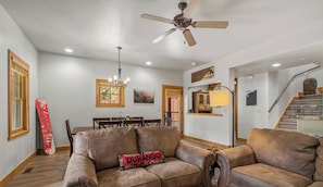 A spacious living room with a brown leather sofa, a ceiling fan, a dining area with wooden chairs, and a staircase.