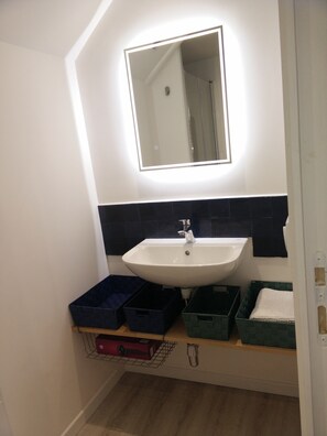 Une salle de bain avec un lavabo blanc, un miroir et des carreaux noirs. On trouve des boîtes de rangement sous le lavabo.