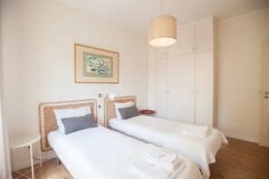 A room with two beds, a small table, a wardrobe, and a hanging light fixture.