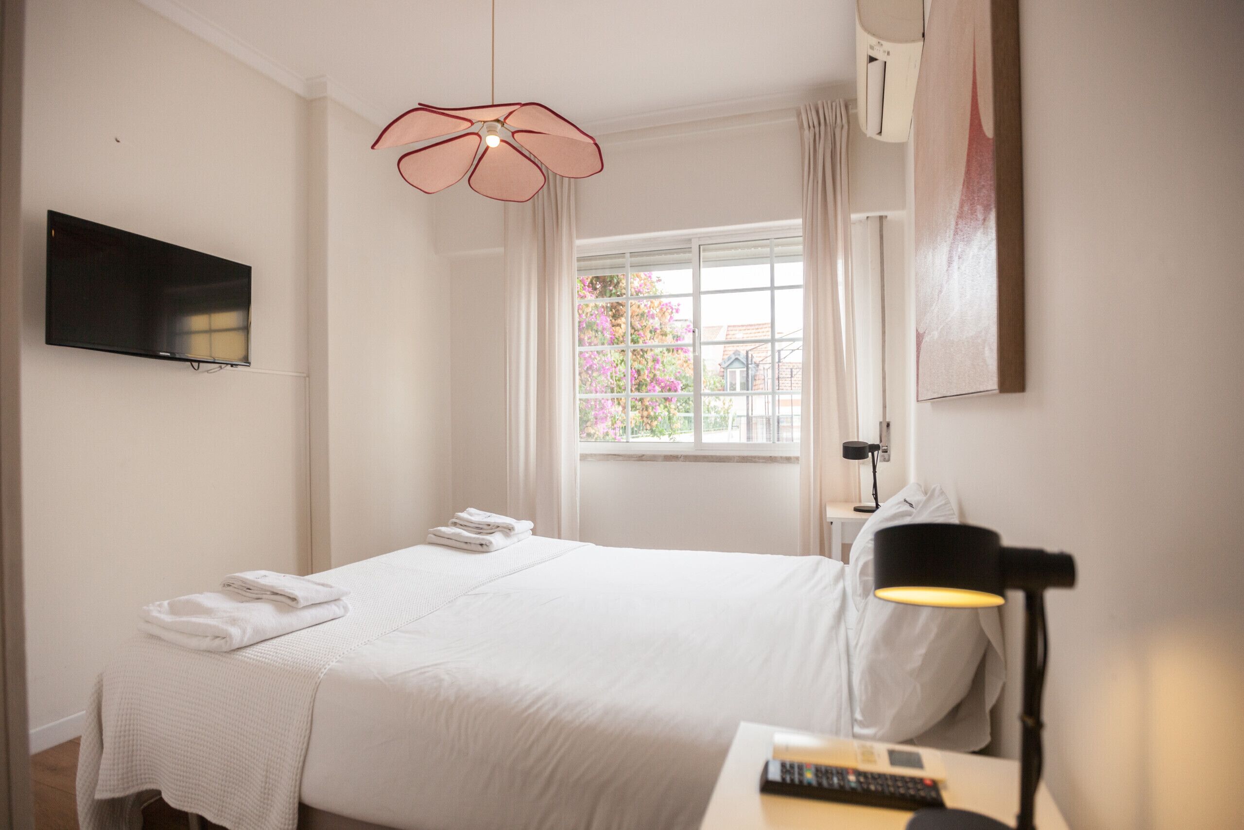 A hotel room with a bed, a flat-screen TV, a window with a view of flowers, and a desk with a lamp and remote control.
