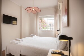 A hotel room with a bed, a flat-screen TV, a window with a view of flowers, and a desk with a lamp and remote control.