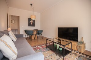 A modern living room with a grey sofa, a coffee table, a flat-screen TV, and a dining area with a round table and chairs.