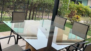 Un patio al aire libre con una mesa y sillas de vidrio, rodeado de follaje y edificios modernos al fondo.