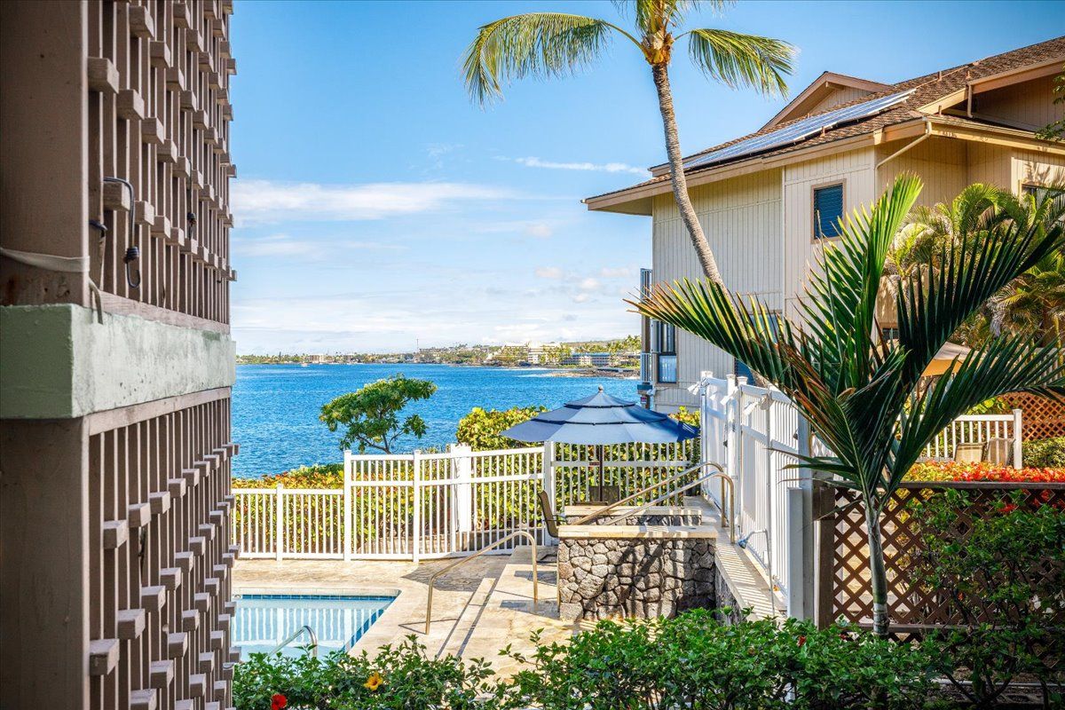 A pool area with a view of the ocean, a white fence, and a palm tree.