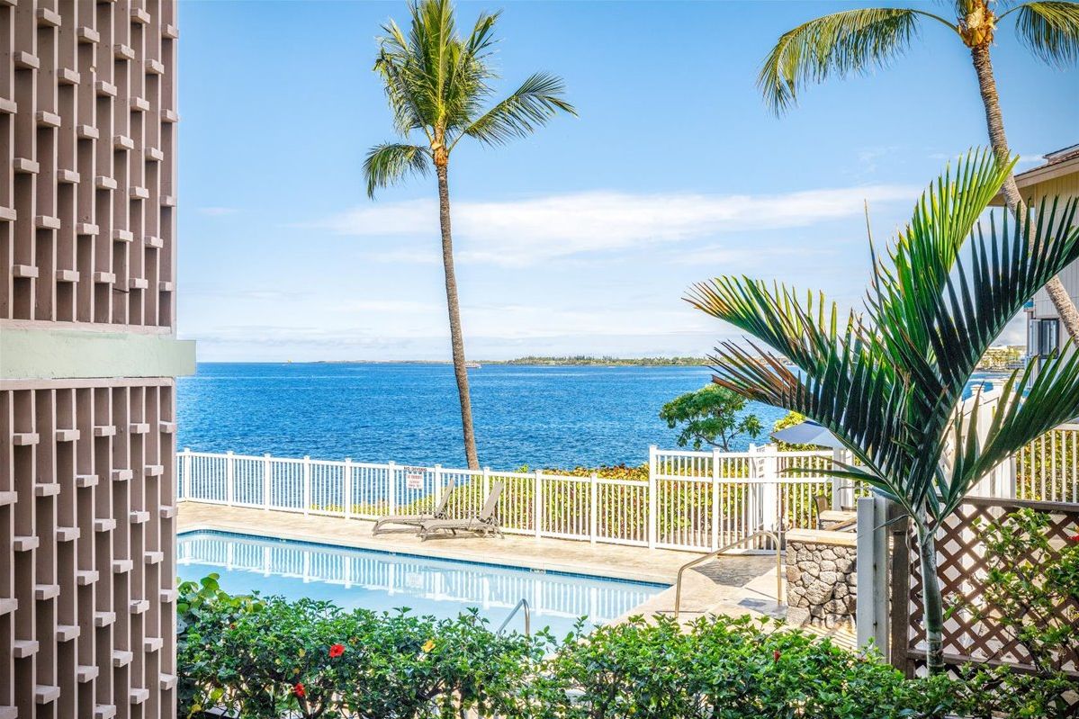 A pool area with a view of the ocean, palm trees, and a white fence.