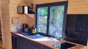 A modern kitchen with wooden cabinetry, a built-in stove, and a large window offering a view of the outdoors. - Roulotte Escapade nature in Morainville (Morainville-Jouveaux)