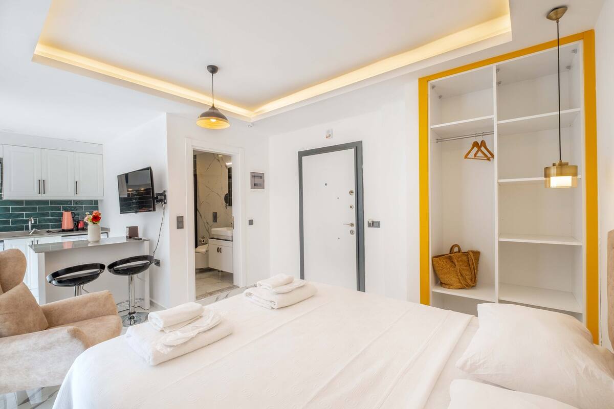 Quarto de hotel com cama, escrivaninha com duas cadeiras, banheiro e guarda-roupa com prateleiras.