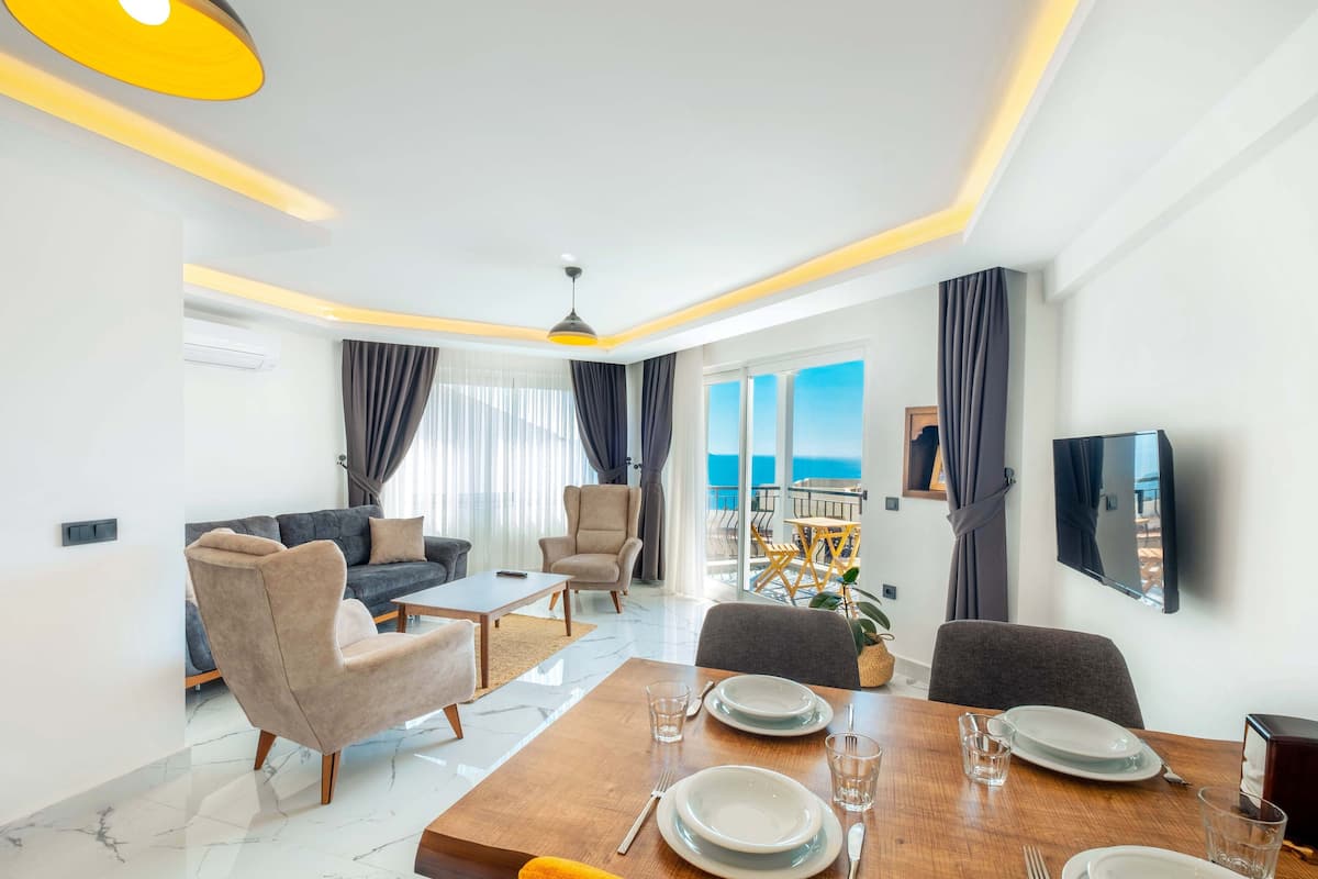 Uma sala de estar moderna com área de jantar, um sofá, uma televisão e uma varanda com vista para o mar.