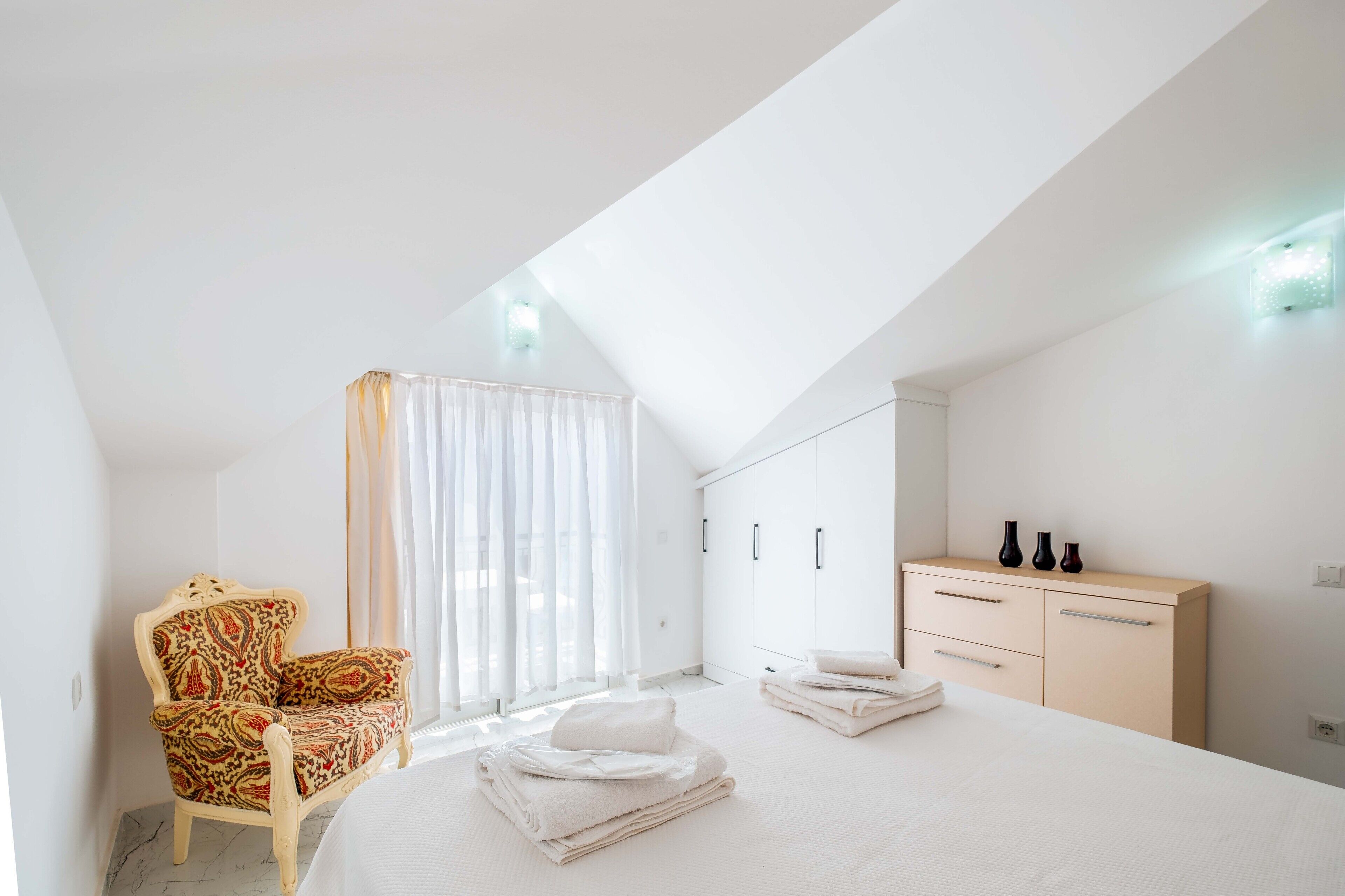 Un dormitorio moderno con una cama grande, una silla, un armario y una ventana con cortinas.