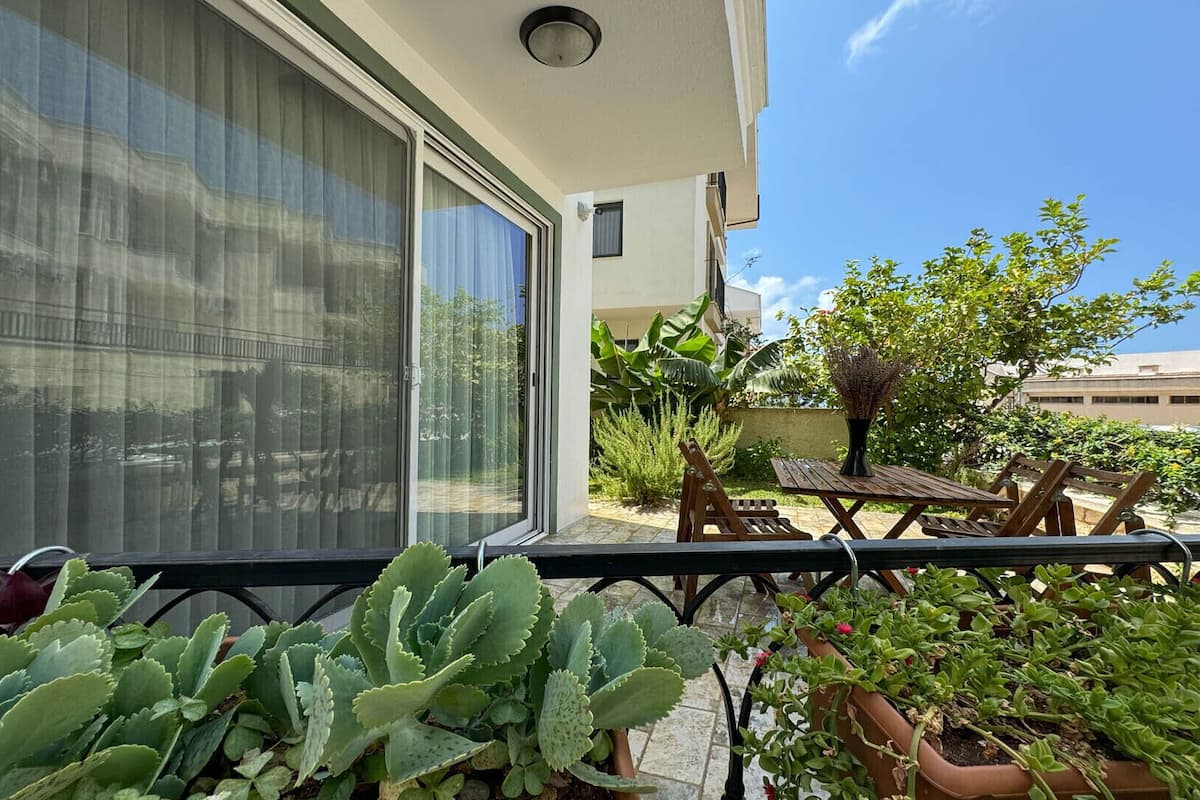 Varanda com mesa e cadeiras, rodeada por plantas em vasos e vista para um edifício.