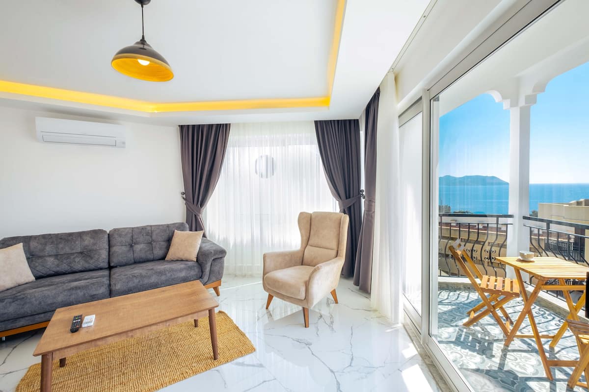 Uma sala de estar moderna com sofá, poltrona e mesa de centro. É visível uma varanda com móveis de exterior através de grandes janelas.