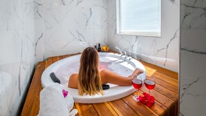 Una mujer relajándose en una bañera con suelo de madera, pared de mármol blanco y una ventana con persianas.