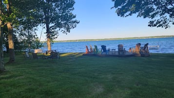 A lakeside deck with picnic tables and Adirondack chairs, surrounded by trees and overlooking a calm lake.